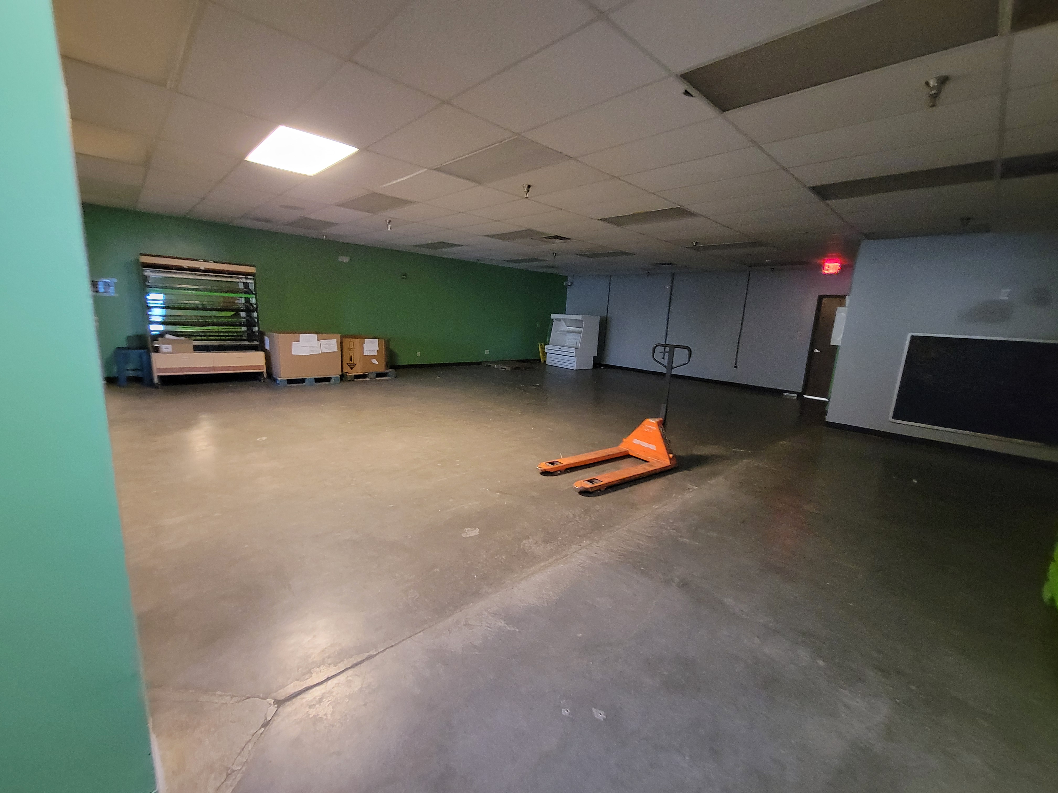 Mostly empty room with pallet jack in the middle of the room plus a fridge unit, shelves, and cardboard containers lined up to the walls.